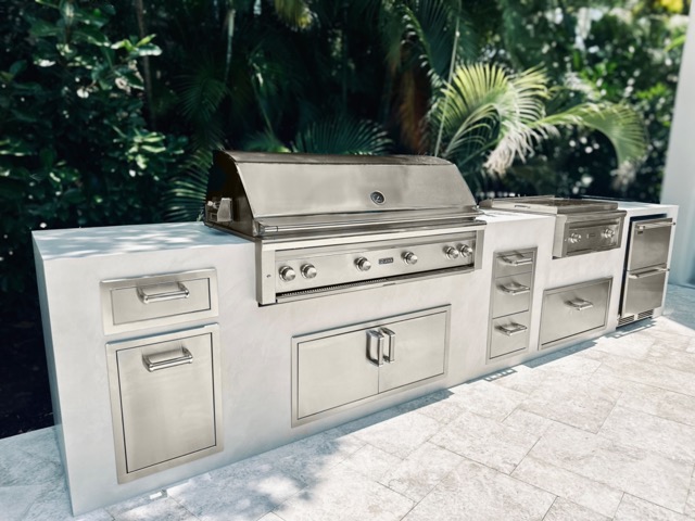 Stainless steel outdoor kitchen with built-in grill, storage cabinets, drawers, and countertop in a tropical outdoor setting.