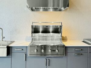 Built-in stainless steel outdoor gas grill with the lid open, installed in a gray outdoor kitchen cabinet beneath a stainless steel vent hood and surrounded by light countertops.