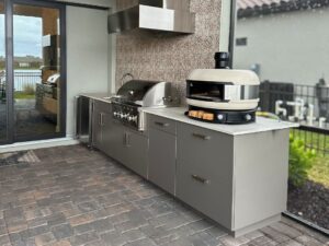Outdoor kitchen installation featuring a built-in gas grill, pizza oven, stainless steel cabinets, and vent hood on a covered patio.