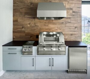 Modern outdoor kitchen with built-in stainless steel gas grill, ventilation hood, cabinetry, and under-counter fridge against a wood-paneled backsplash.