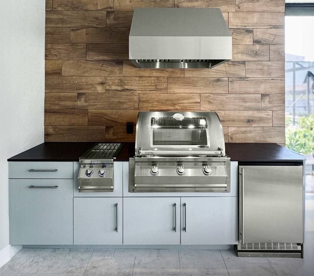 Modern outdoor kitchen with built-in stainless steel gas grill, ventilation hood, cabinetry, and under-counter fridge against a wood-paneled backsplash.