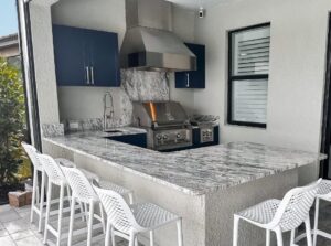 Outdoor kitchen with a large white and gray quartz countertop, built-in stainless steel gas grill and range hood, blue cabinetry, bar seating, and a matching quartz backsplash in a covered patio.