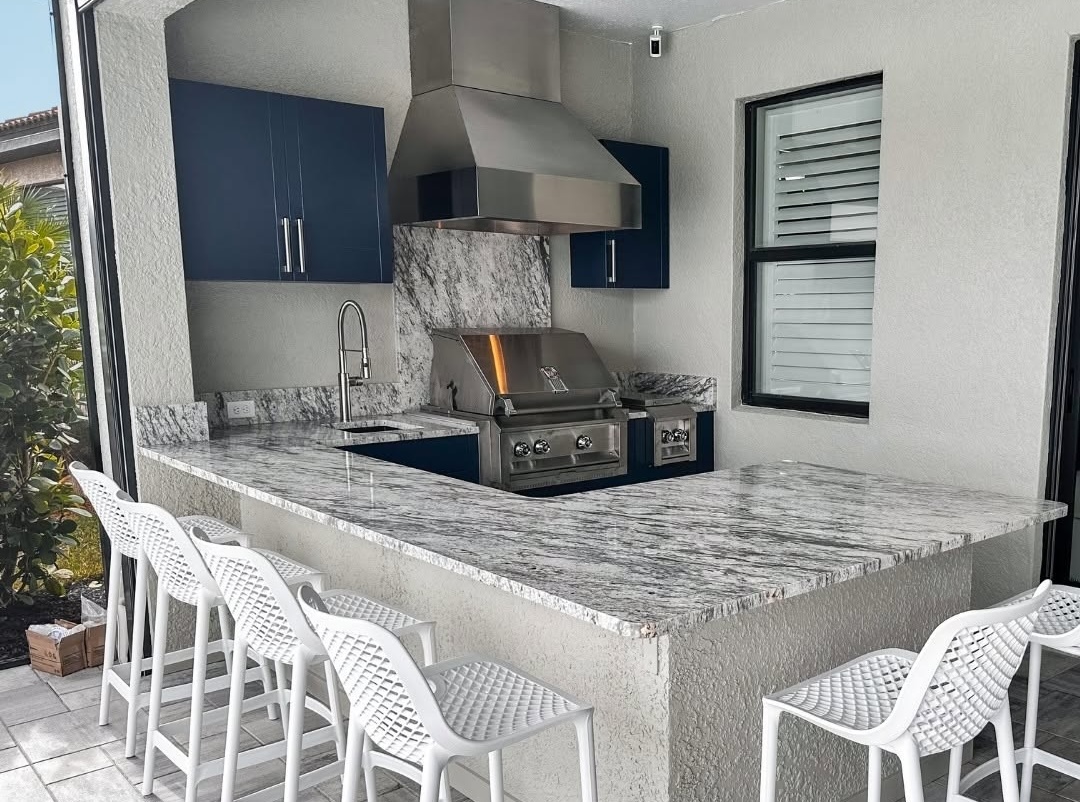 Outdoor kitchen with a large white and gray quartz countertop, built-in stainless steel gas grill and range hood, blue cabinetry, bar seating, and a matching quartz backsplash in a covered patio.