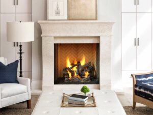 A traditional fireplace with a lit fire in a bright, elegant living room with white cabinetry, armchairs, and a coffee table.